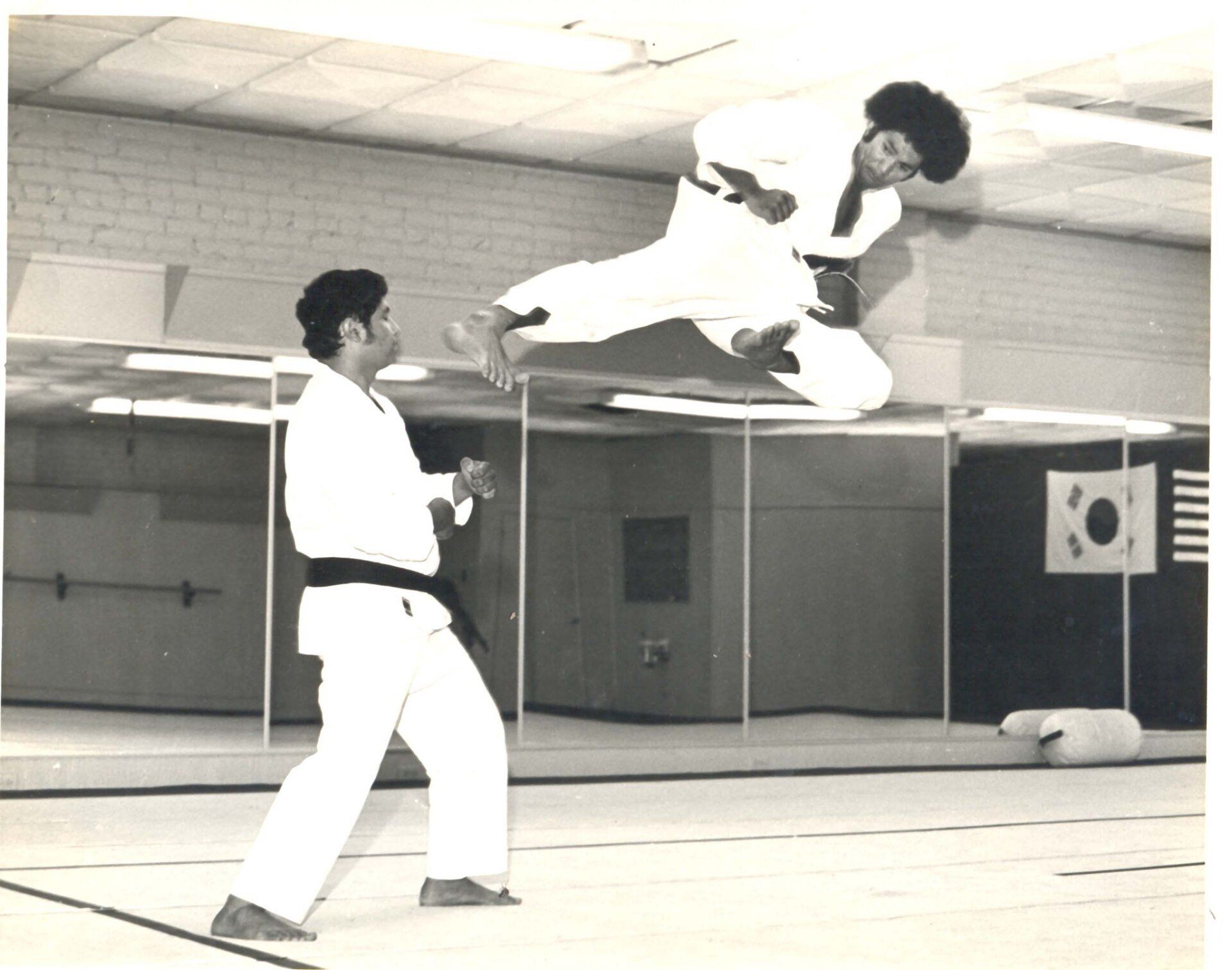 Two men in karate uniforms practicing in a gym. One performs a jumping kick, while the other stands ready.