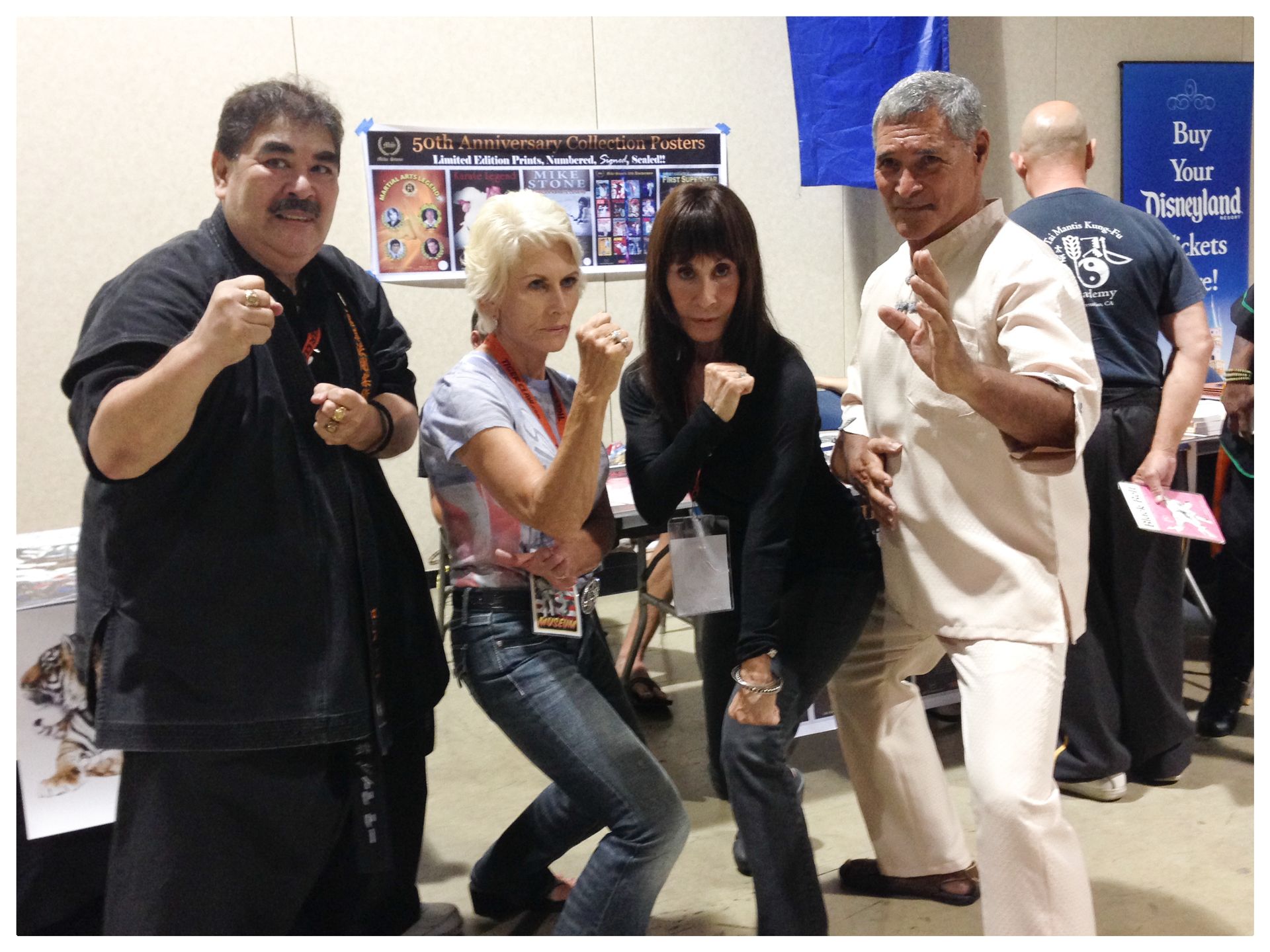 Four people posing, fists raised, in a martial arts stance at a convention booth. They are smiling and looking at the camera.