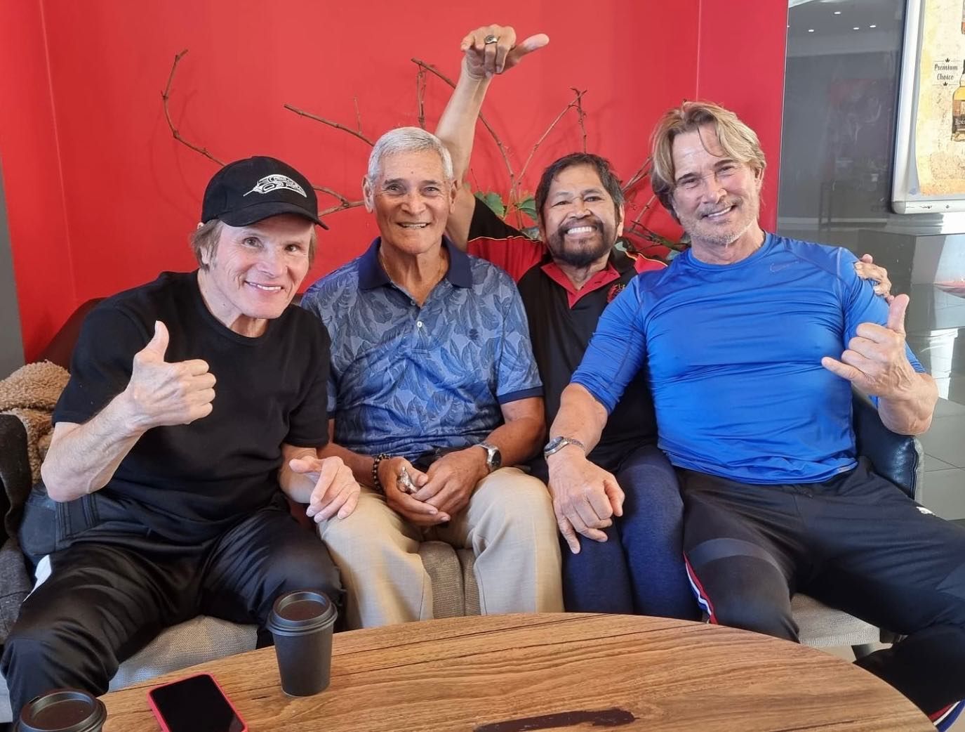 Four men sitting on a couch, one raising a hand with fingers extended. They are smiling, the setting has a red wall.