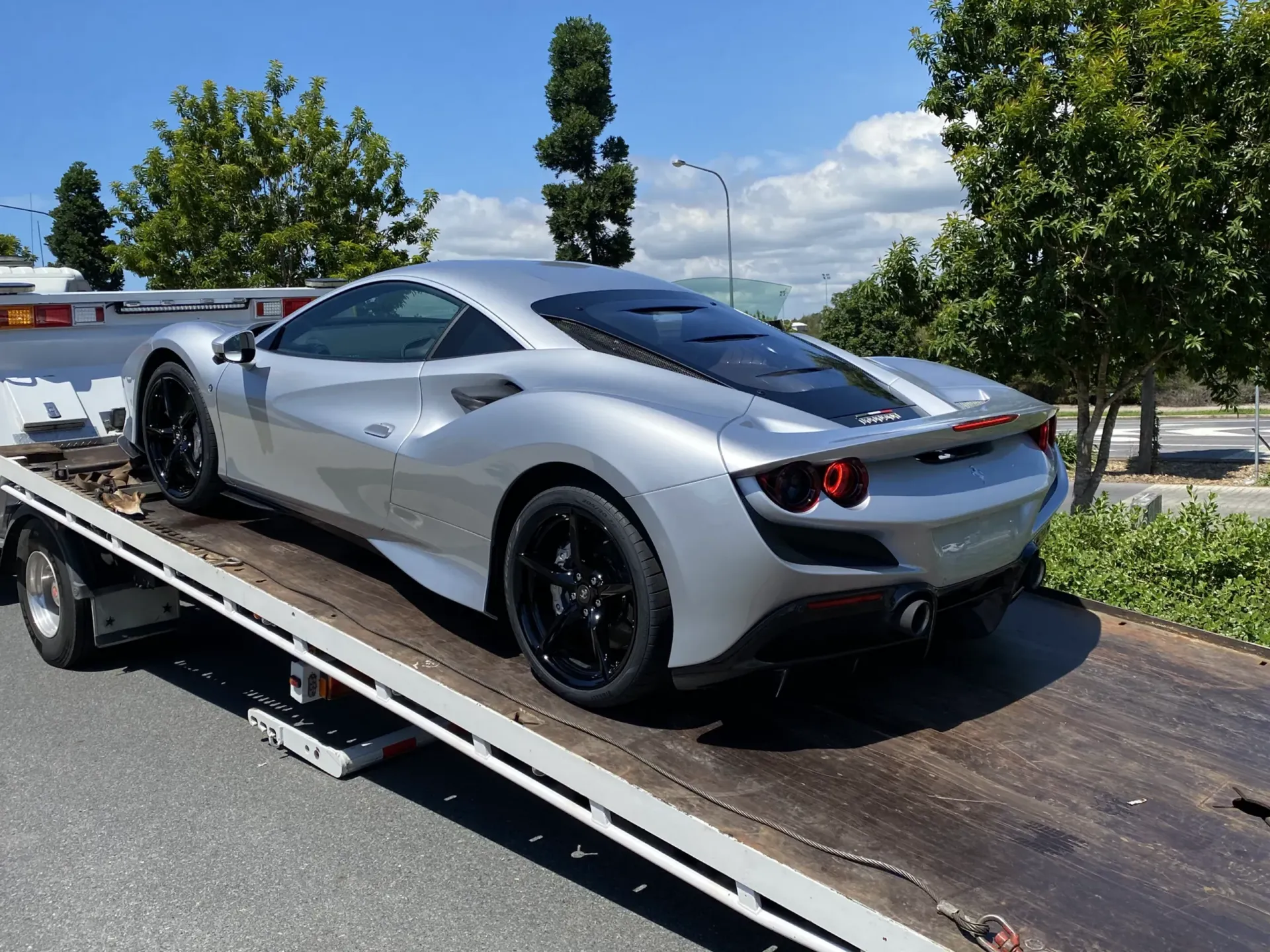 Silver Ferrari sports car on a flatbed tow truck. Black wheels, sunny outdoor setting.