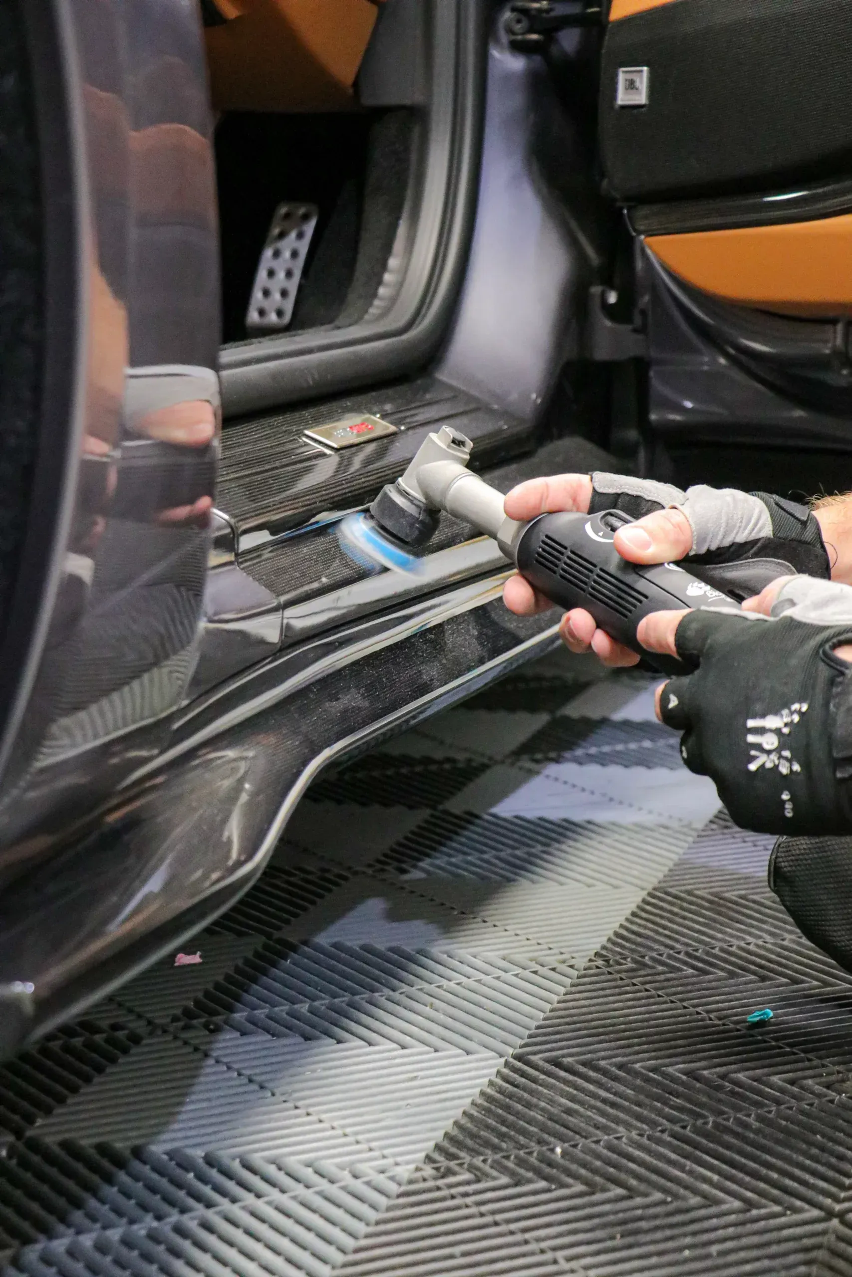 Person cleaning car's side skirt with a power tool; interior of car visible; gray and black color scheme.