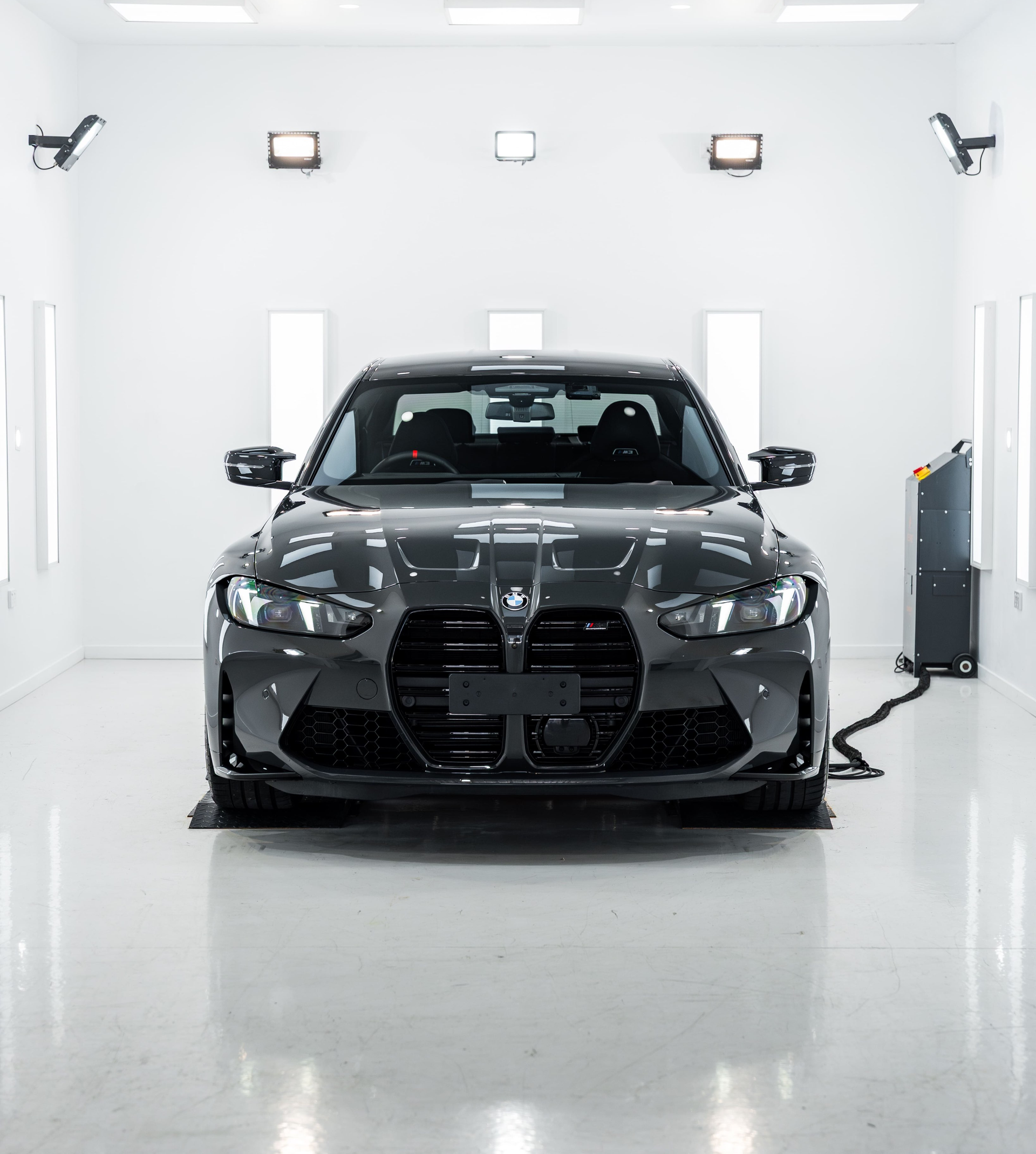 A dark-colored BMW car parked in the center of a brightly lit, sterile white studio with reflective floors.