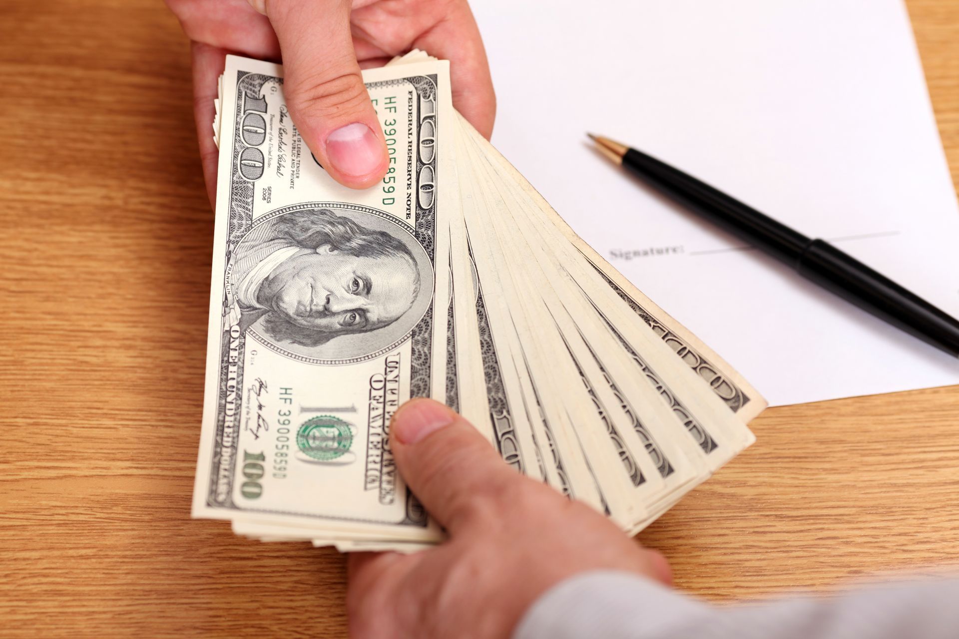 Hands exchanging U.S. dollar bills over a wooden desk beside a pencil and paper