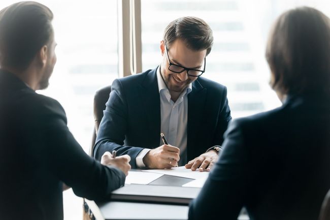 Three people in a bright office meeting, reviewing documents at a table.