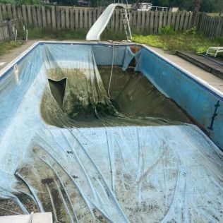 Empty, neglected swimming pool with a dirty liner, slide, and fence in the background.