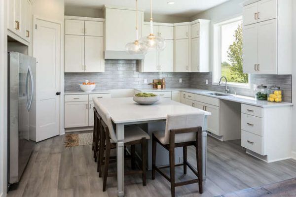 White kitchen with island, cabinets, and wood-look floor. Light gray backsplash, stainless steel fridge, and stools.