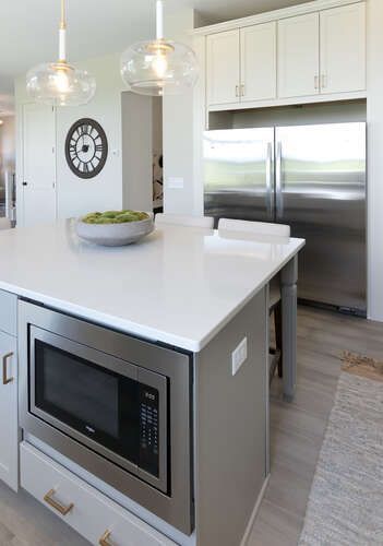 Kitchen island with built-in microwave, white countertop, light gray cabinets, and stainless steel refrigerator.