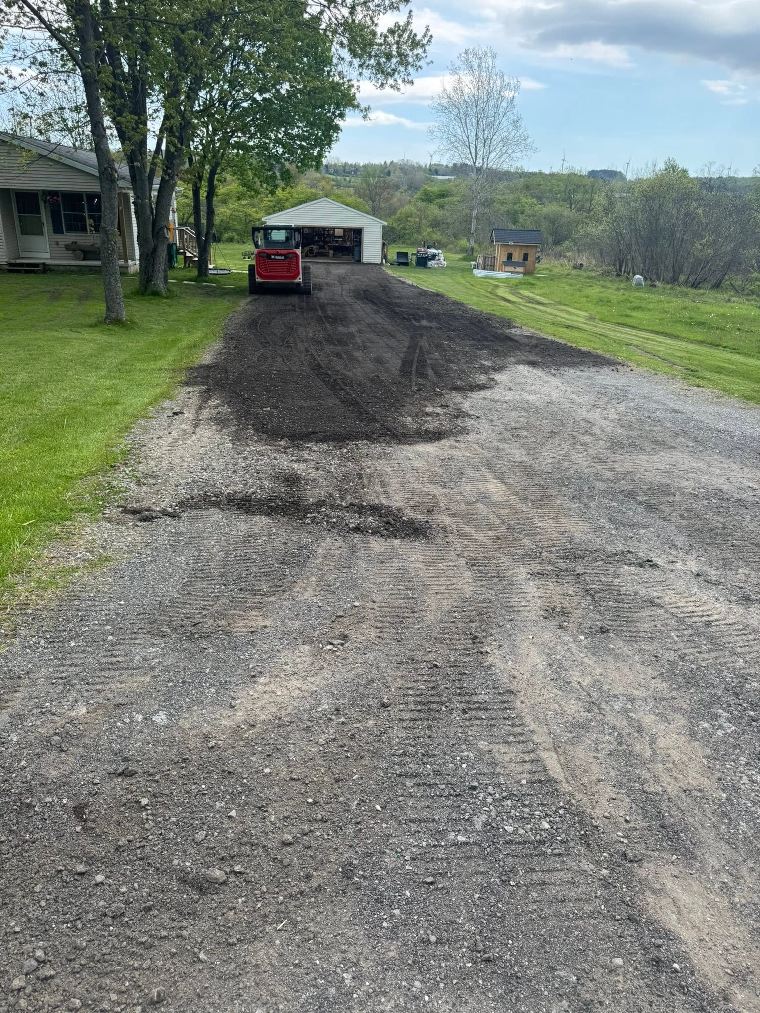 Gravel driveway with a red lawn mower, muddy dirt pile, trees, and a house in the background