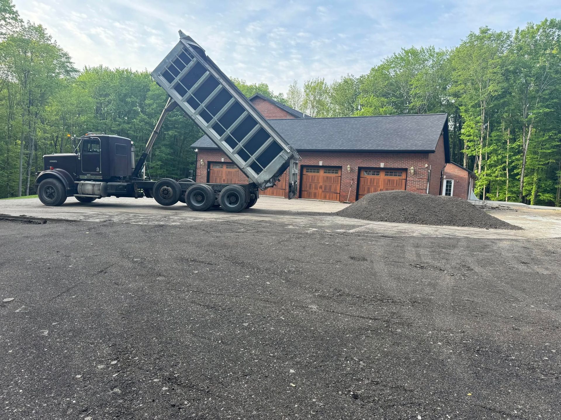 Dump truck raising bed to unload gravel beside a brick building and wooded area.