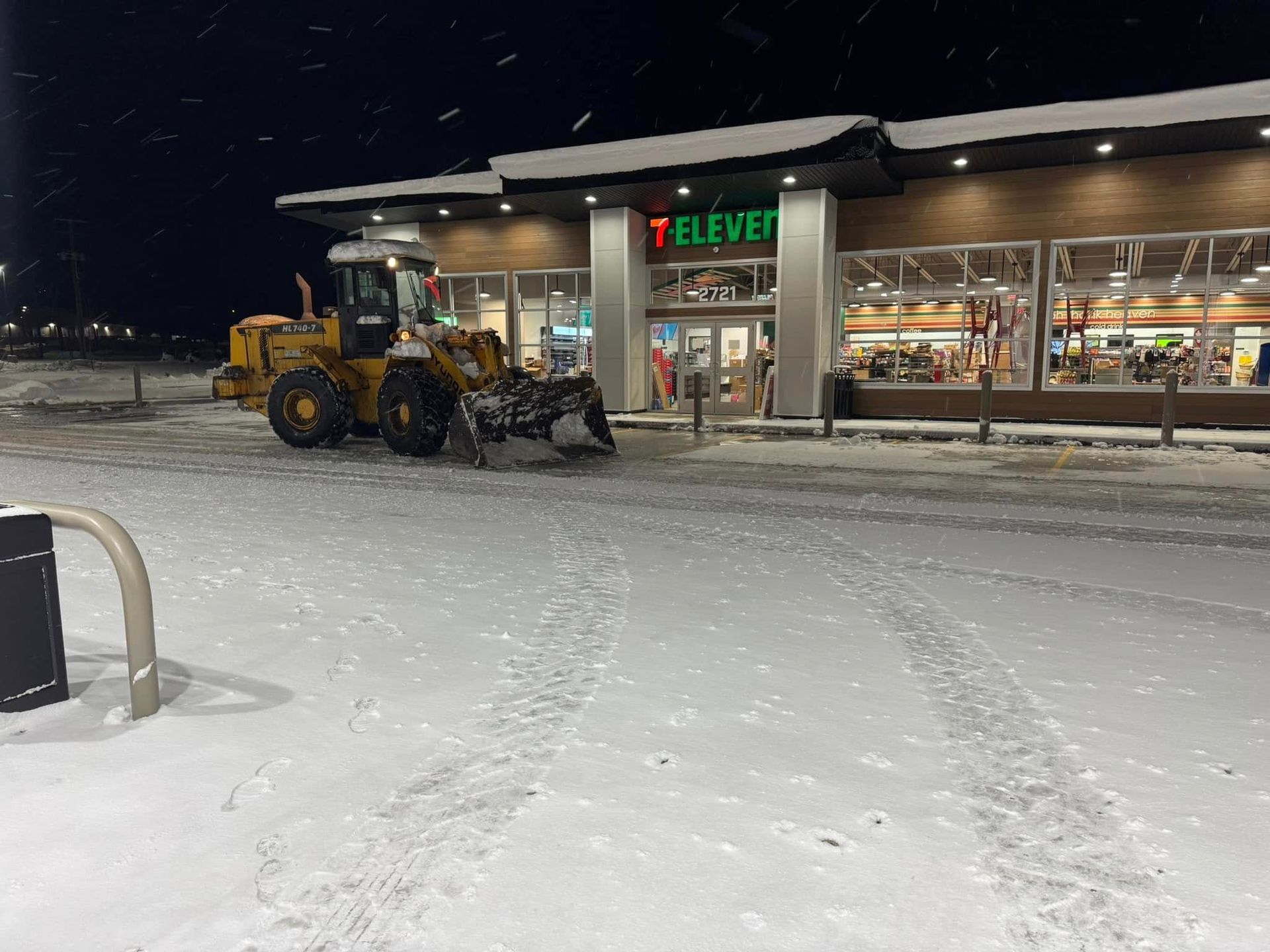 Snowy 7-Eleven parking lot at night with a yellow loader clearing snow near the entrance
