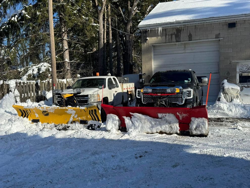 Two snowplows, one yellow and one red, clearing snow in a driveway beside a building.