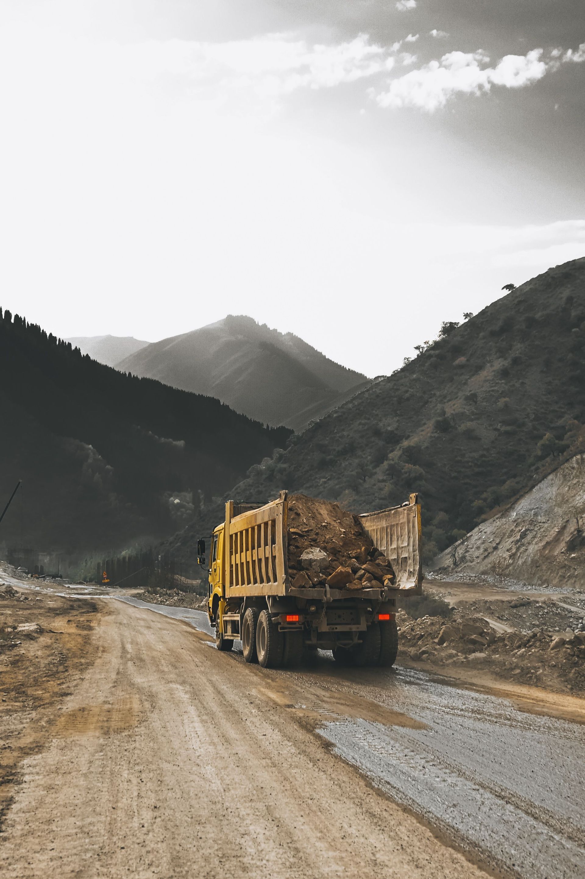 Yellow dump truck on a muddy mountain road with towering rocky hills in the background