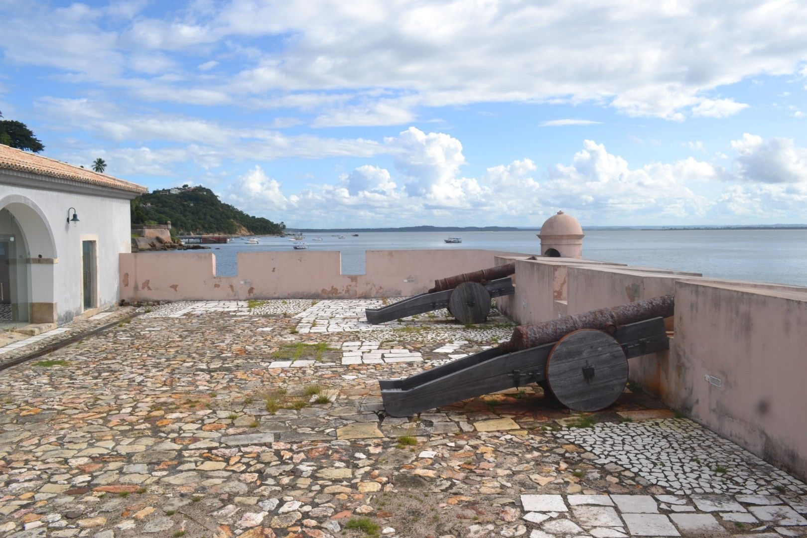 Prédio rosa ao lado de uma rua asfaltada com carros e árvores, sob um céu azul.