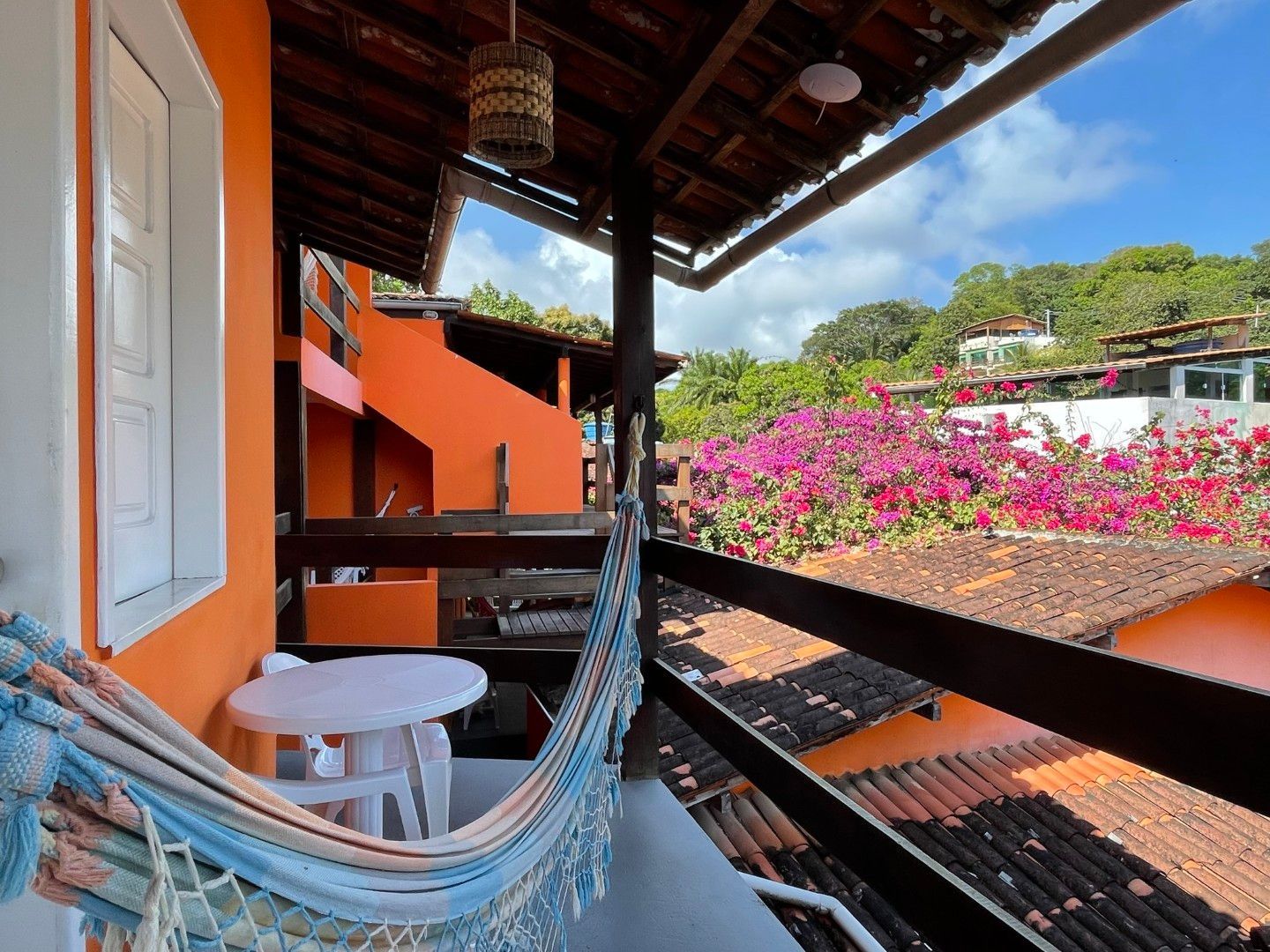 Rede numa varanda sombreada com vista para paredes alaranjadas, flores cor-de-rosa e um jardim ensolarado na encosta.