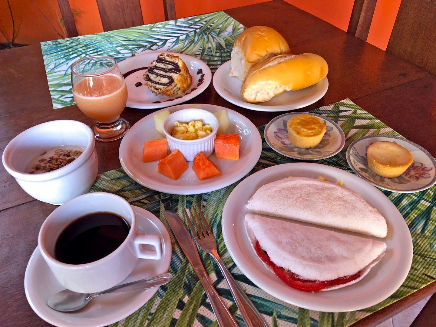 Café da manhã servido em uma mesa de madeira com café, pãezinhos, doces, frutas e pequenos recipientes com geleia e iogurte.