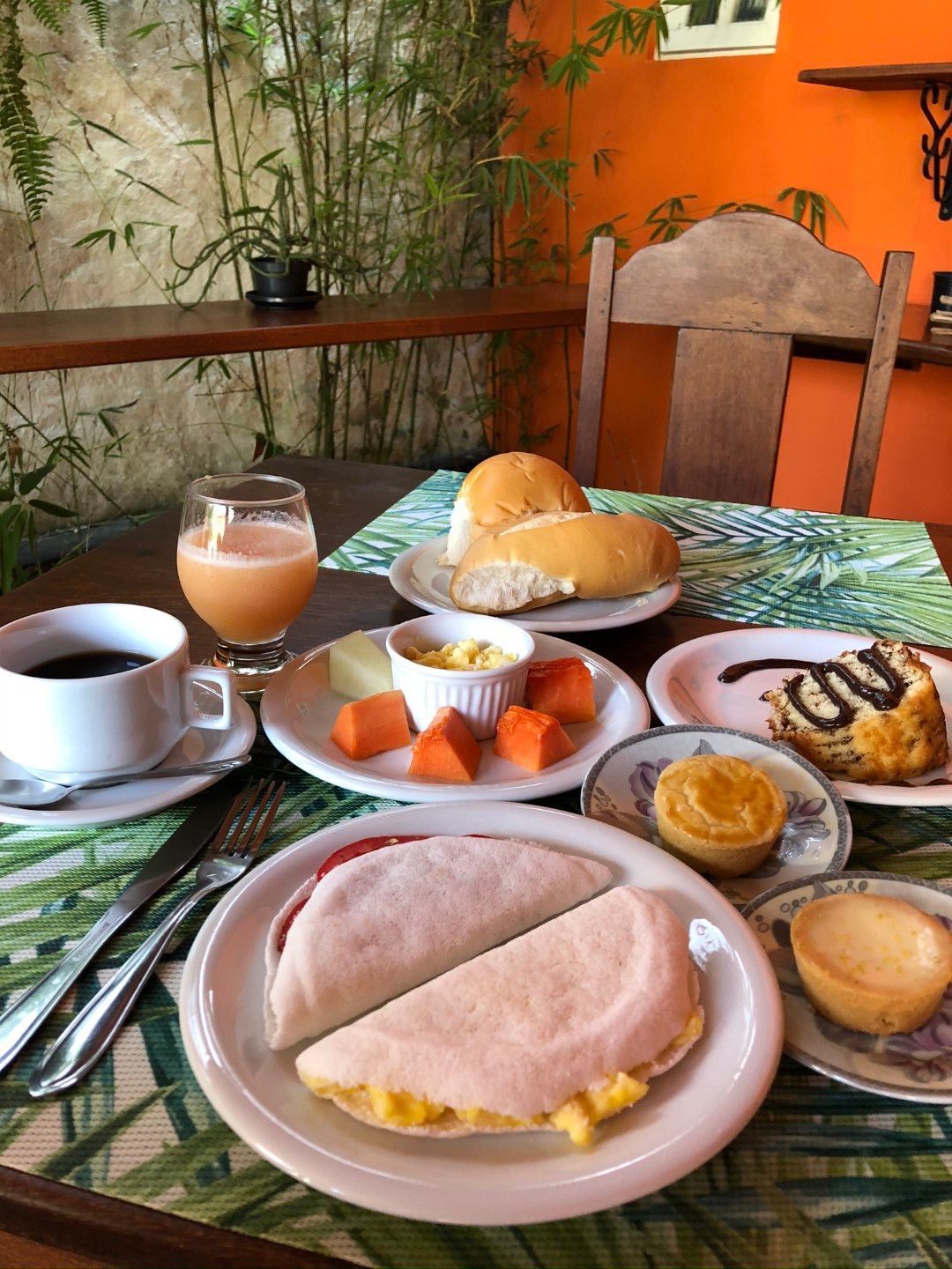 Mesa de café da manhã com café, chá, doces, frutas e um sanduíche em um ambiente aconchegante de café.