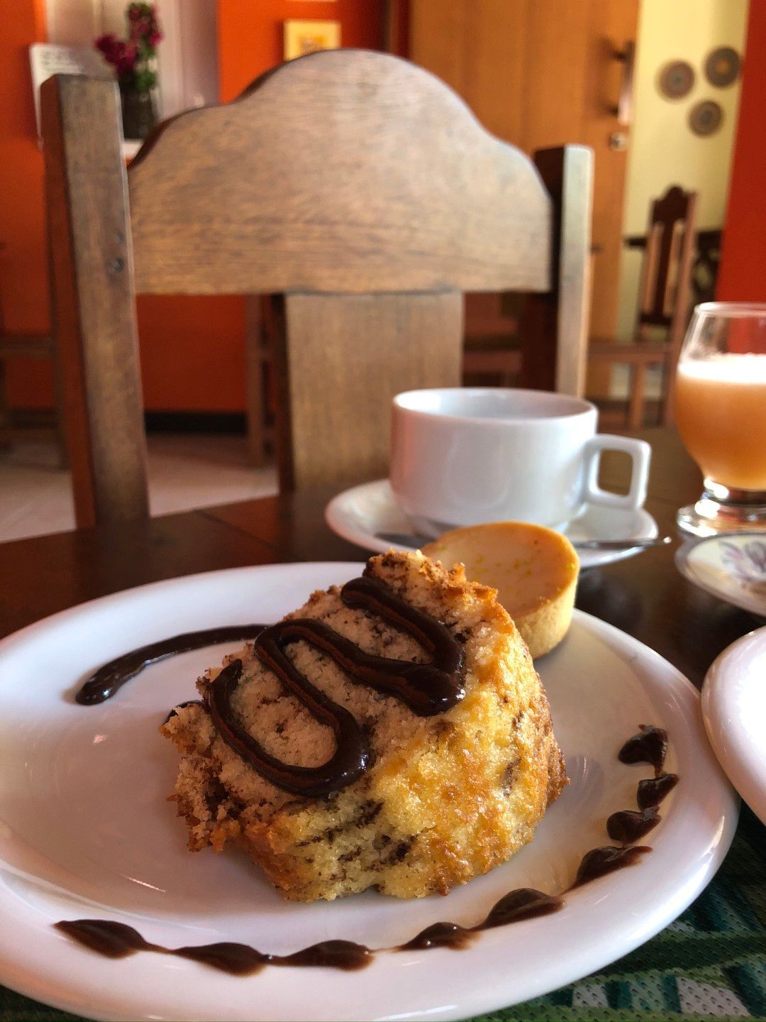 Pastel com cobertura de chocolate servido em um prato branco, acompanhado de café, em um ambiente aconchegante de cafeteria.