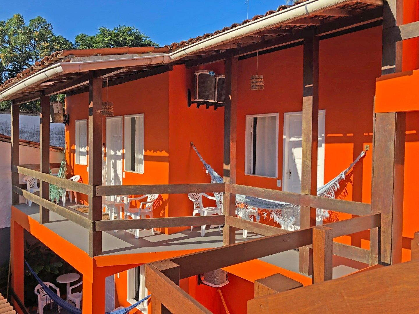 Varanda de casa laranja com portas brancas, corrimão de madeira e rede sob a luz do sol.