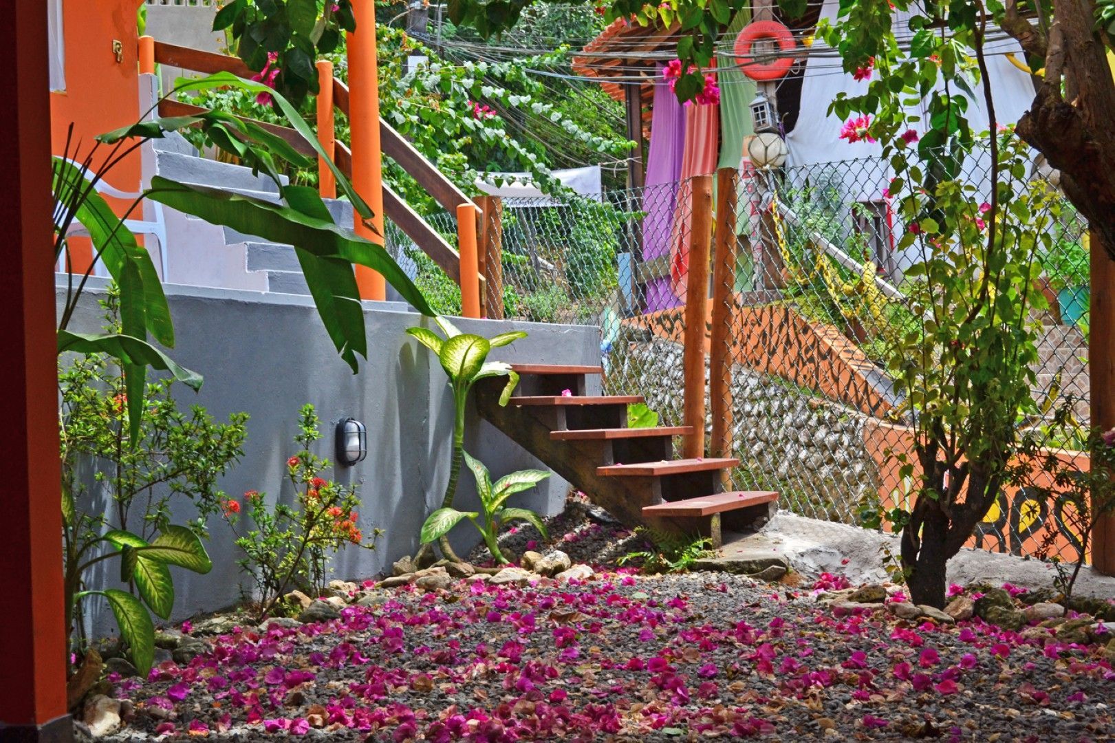 Escadaria de jardim com flores cor-de-rosa, vegetação exuberante e fitas penduradas ao lado de uma parede cinza.