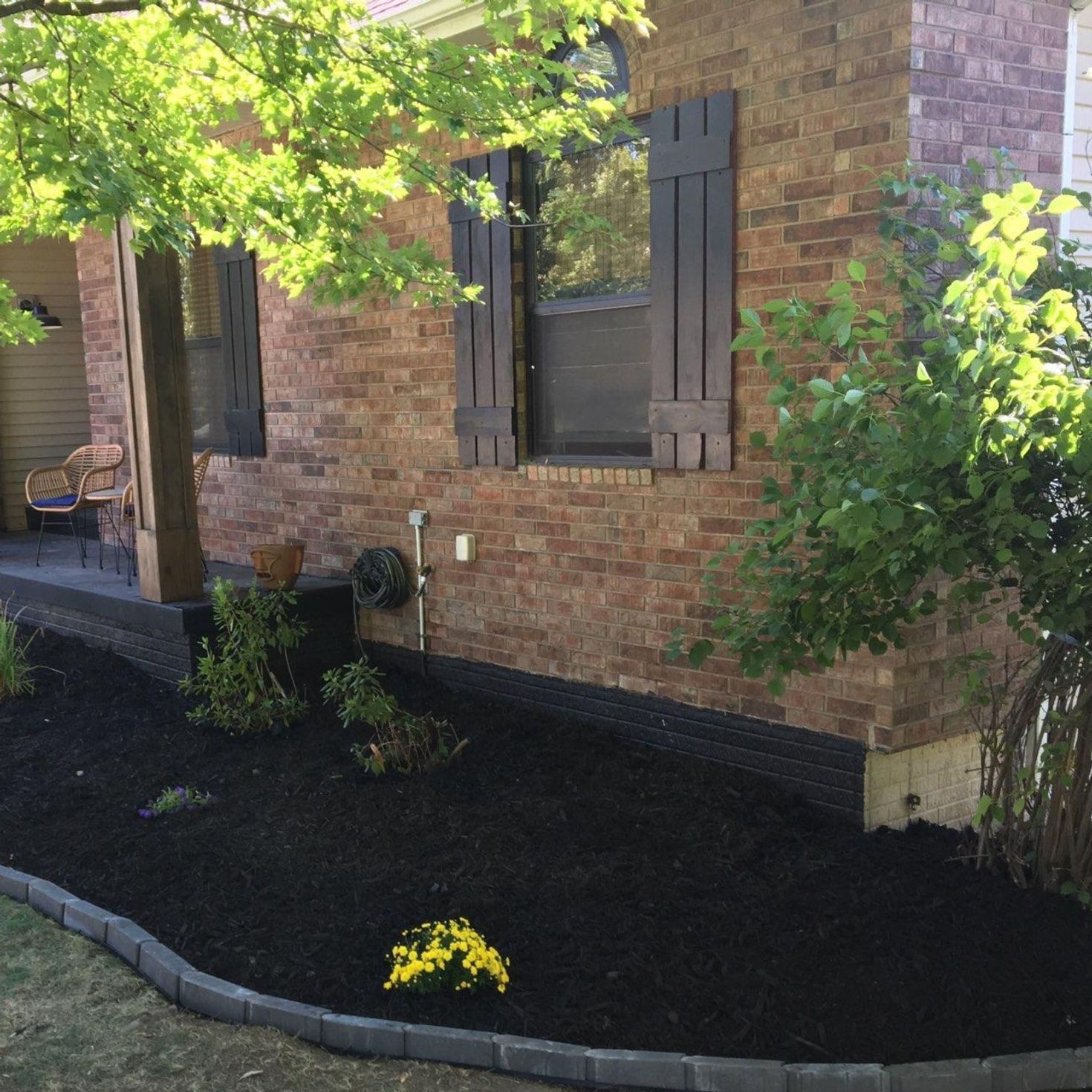 Brick house exterior with dark mulch landscaping, shrubs, and a small yellow flower bed.