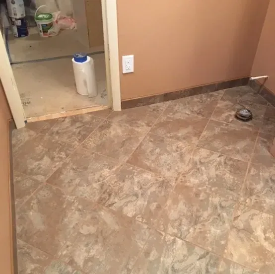 Bathroom floor with tan tile, a doorway to a utility area, and a small pet bowl near the wall.