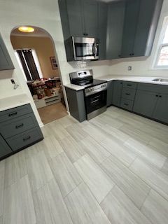 Modern kitchen with gray cabinets, white countertops, and light wood flooring, viewed toward an arched doorway.
