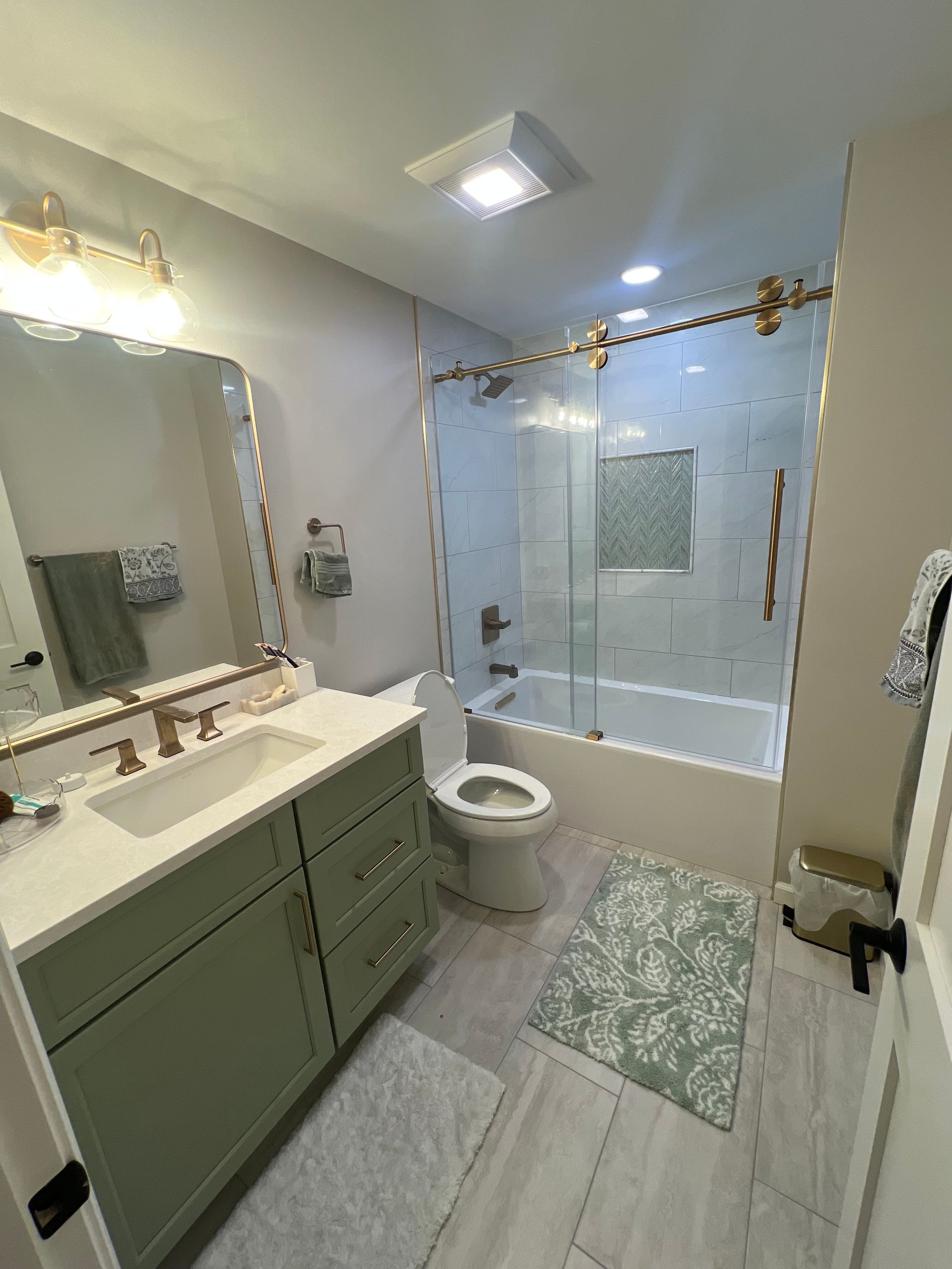 Bright bathroom with vanity, toilet, glass shower, and green cabinet, lit by ceiling lights.
