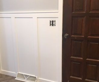 White paneled wall with an outlet and dark wooden door beside a floor vent