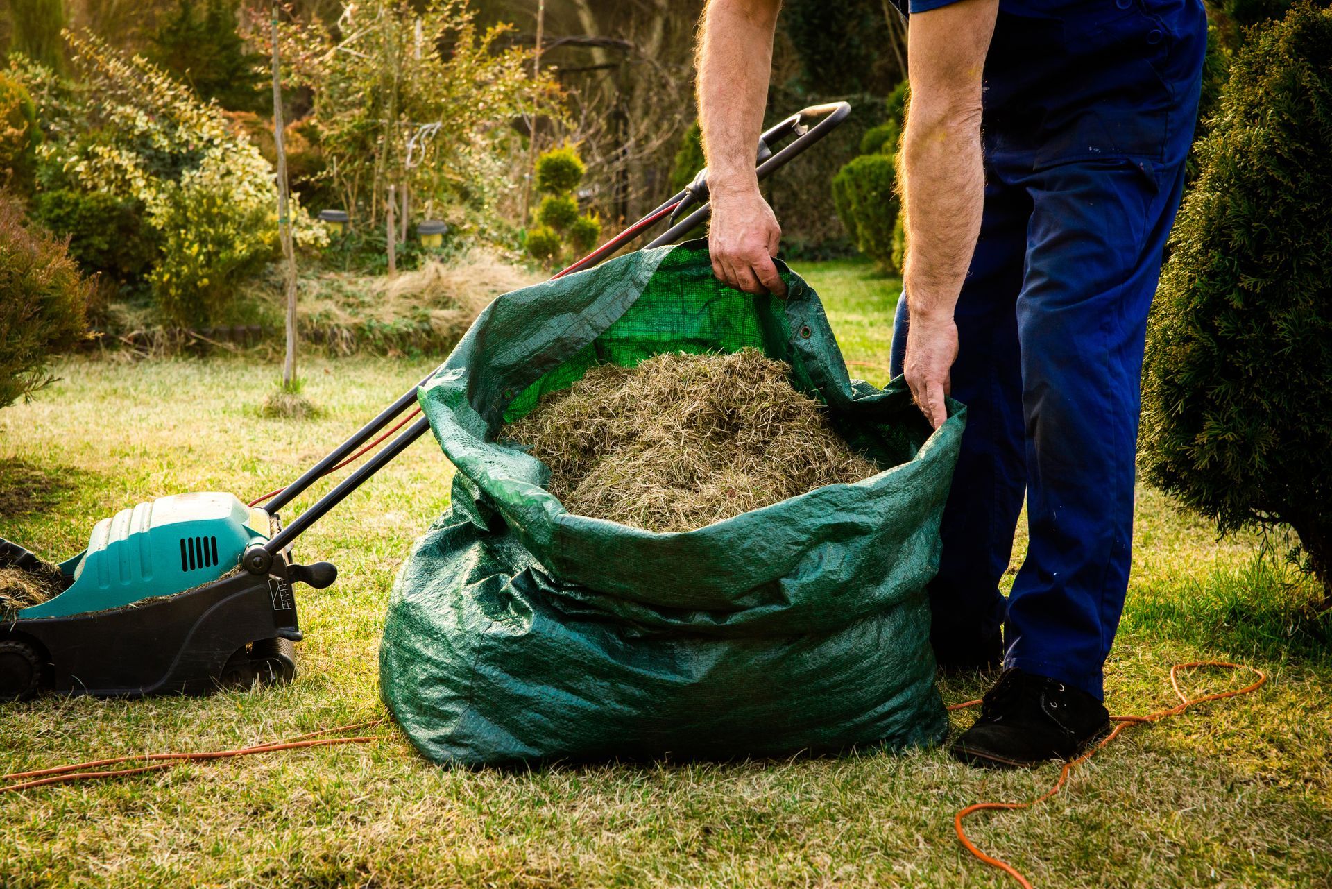 Person emptying grass clippings from a green bag into a lawn. Lawnmower nearby.