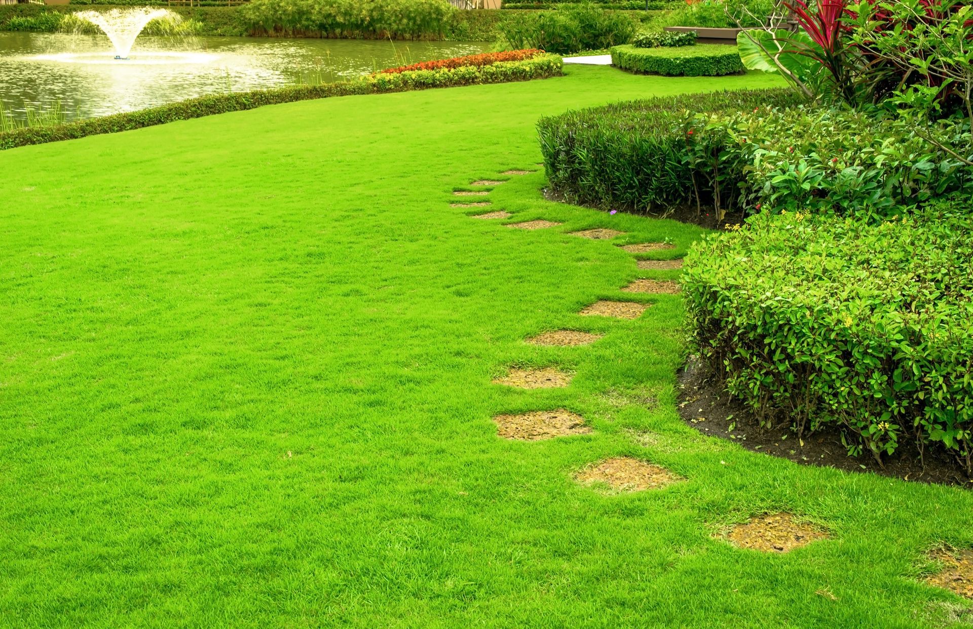 Green lawn with a stone pathway, bushes, and a pond with a fountain.