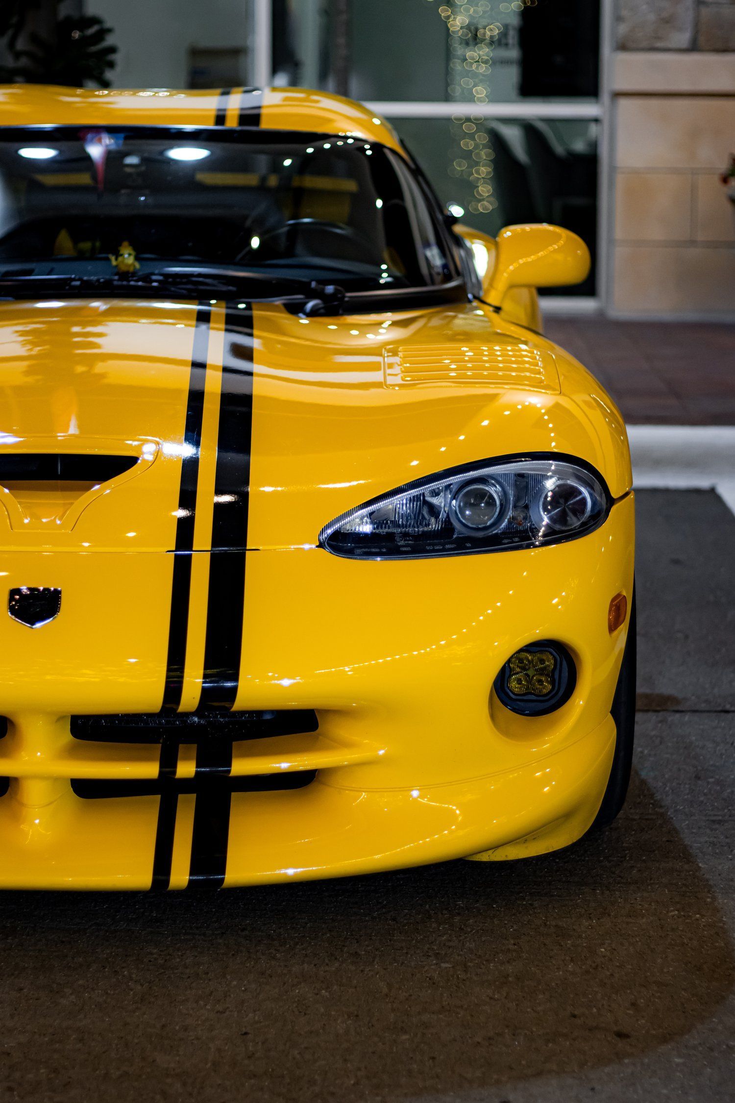 A yellow sports car with black stripes on the hood is parked in front of a building.