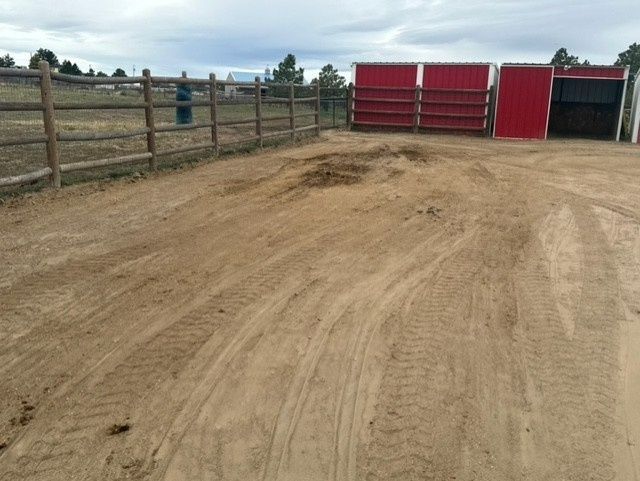 Dusty dirt corral with red barns and a fenced pasture under a cloudy sky