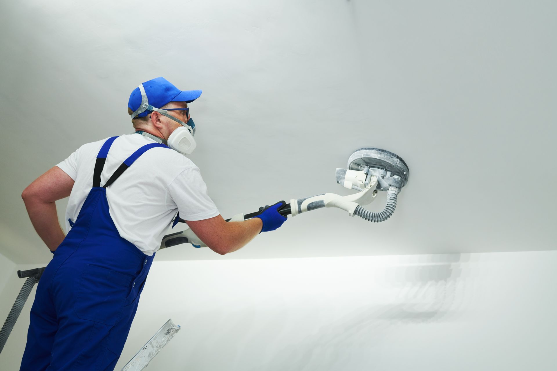 A man wearing a mask is sanding the ceiling with a sander.