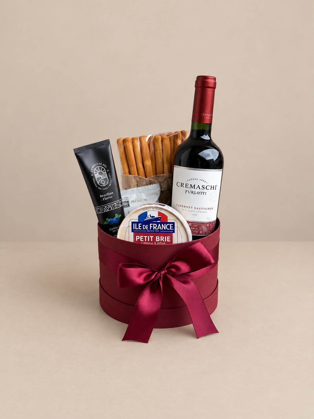 Cesta de presentes com vinho, biscoitos e petiscos, amarrada com uma fita bordô