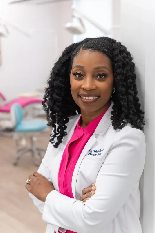Woman in a white coat and pink blouse, smiling with arms crossed in a clinic office.