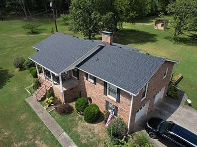 Aerial view of a brick house with a gray roof, front porch, and a driveway in a green yard.