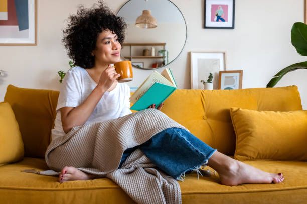 Person sitting on a yellow couch, sipping tea and reading a book in a cozy living room.