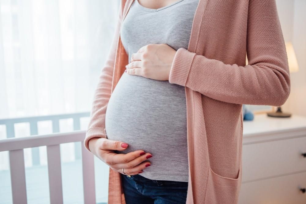 Pregnant person holding belly in a pink cardigan beside a nursery crib