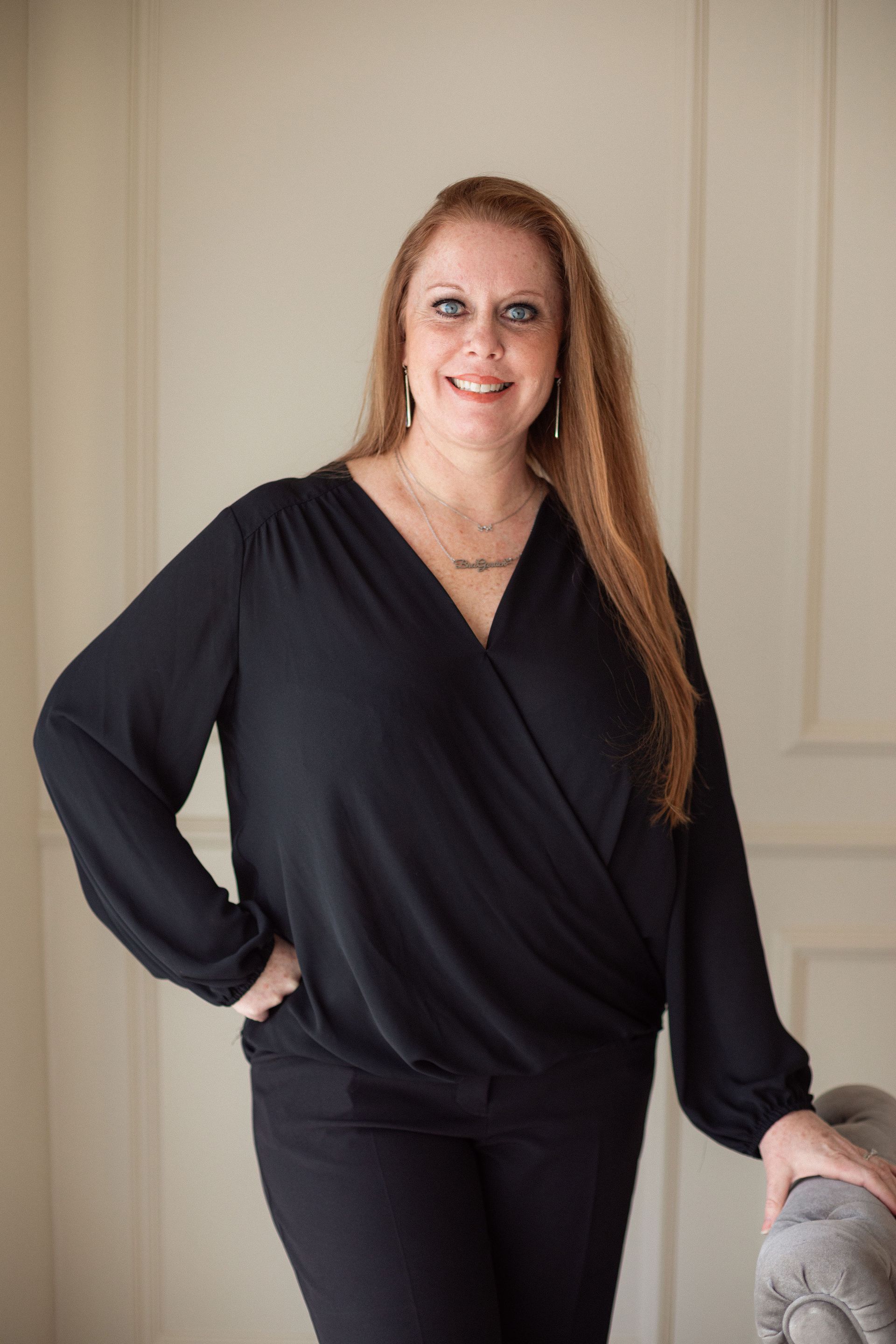 Woman in a black outfit standing by a beige wall, smiling with one hand on her hip.