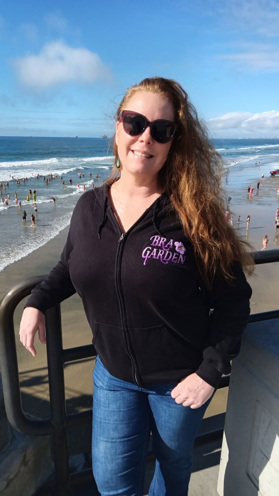 Smiling woman in sunglasses standing by a beach railing with ocean waves and blue sky behind her