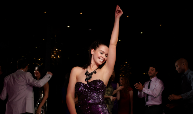 Smiling woman in a purple dress dancing with one arm raised at a dimly lit party