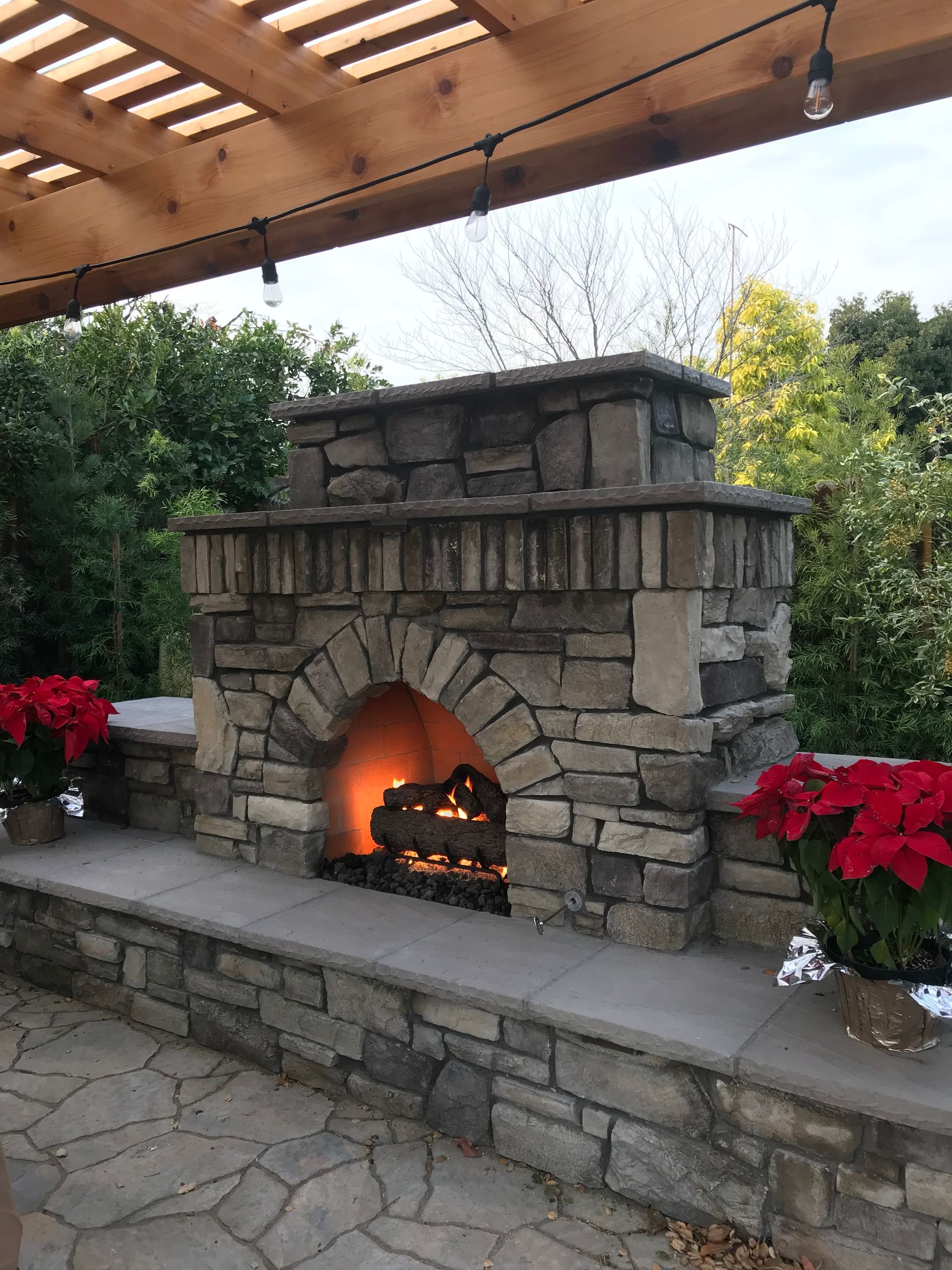 A stone fireplace is sitting under a wooden pergola on a patio.