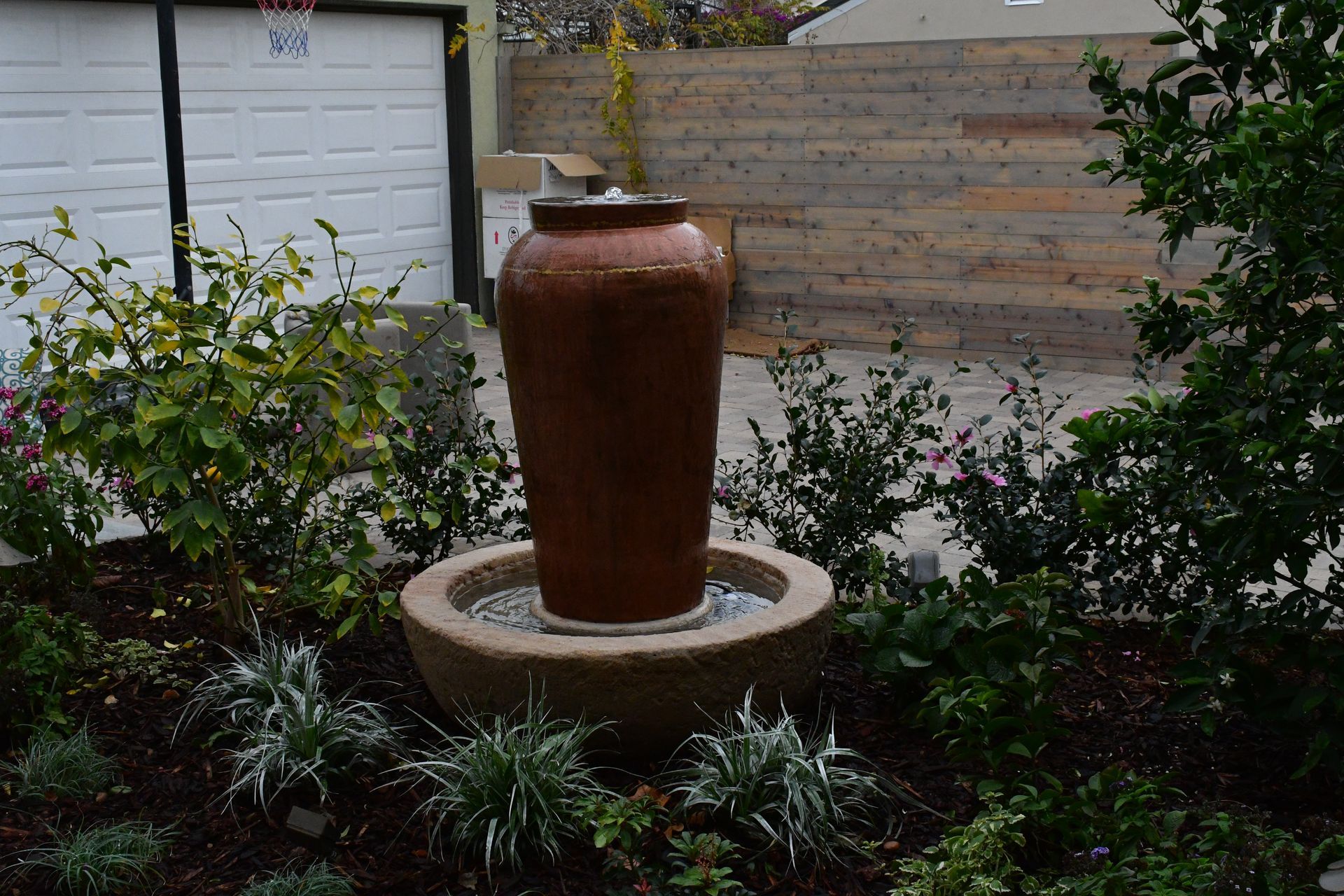 A large vase is sitting in the middle of a garden next to a garage door.