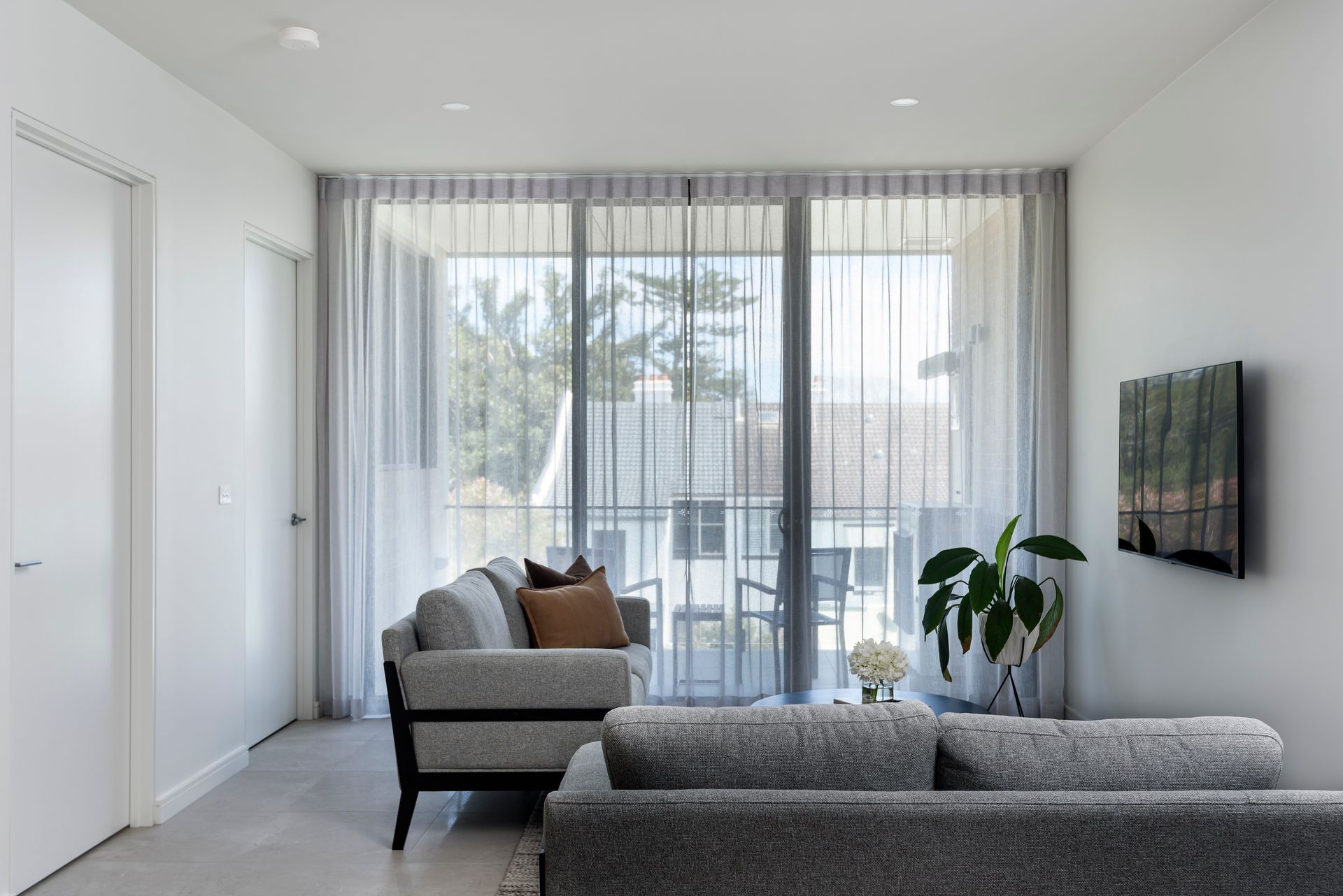 Bright modern living room with gray sofas, sheer curtains, and a wall-mounted TV by a large window.