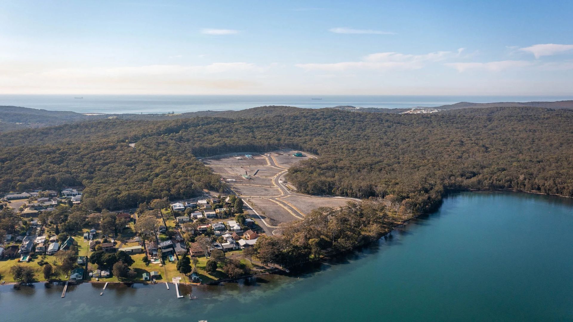 Aerial view of a forested shoreline with a small town, a cleared area, and a blue lake under a clear sky