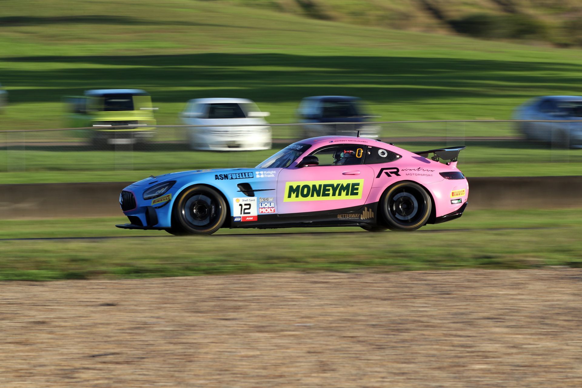Pink and blue race car speeding on a dirt track, with blurred cars and grassy hills in the background