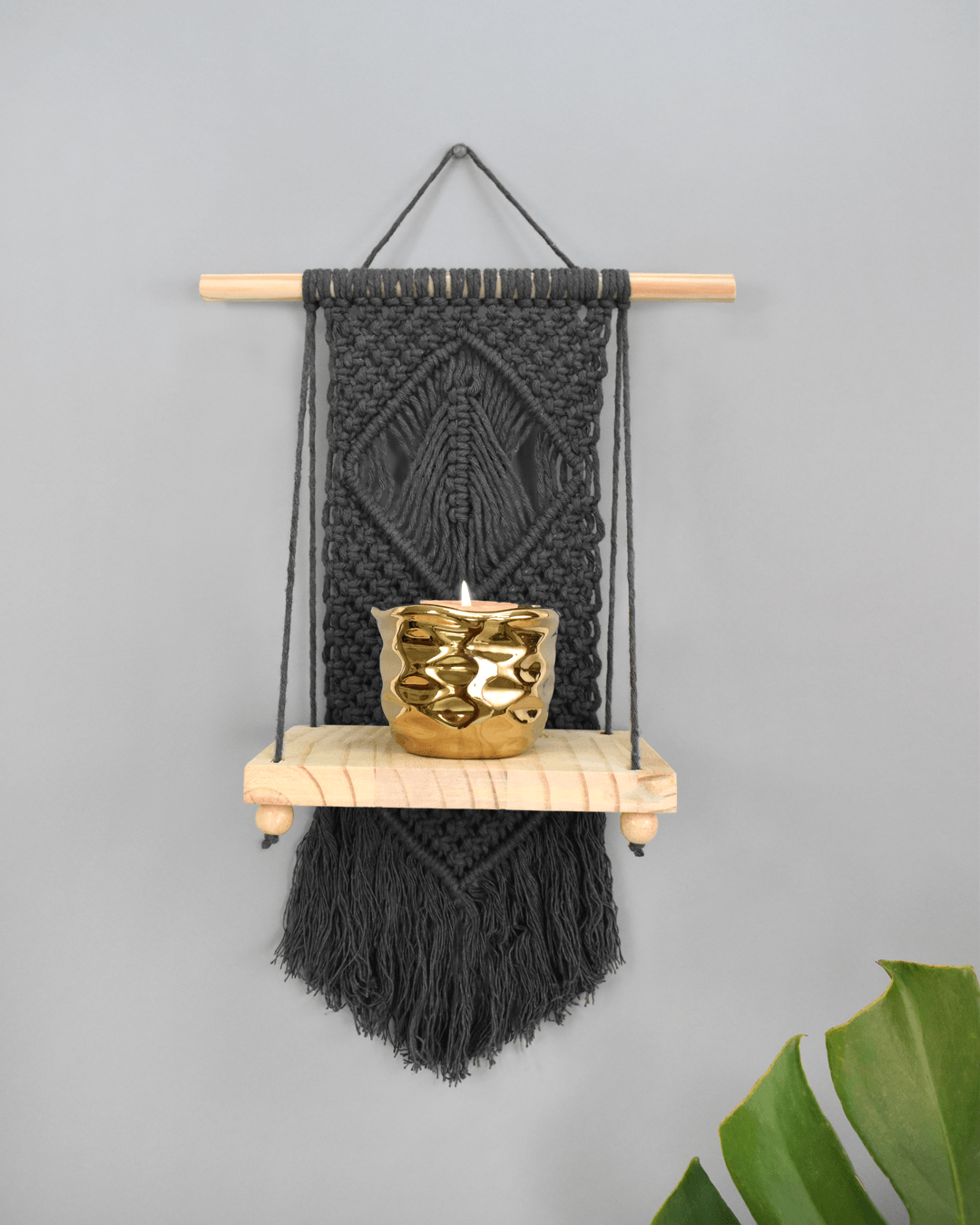 Black macramé wall hanging with wooden shelf and gold candle holder on a gray wall, with a green leaf at right.
