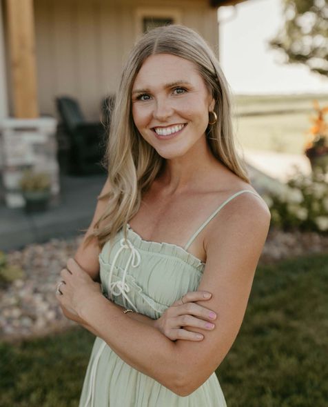 Smiling woman in a light green dress standing outdoors with arms crossed, near a house at sunset