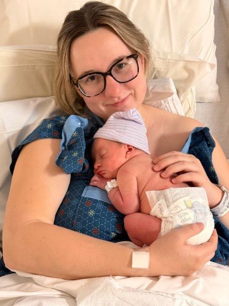 Woman cradling a sleeping newborn in a hospital bed, wearing glasses and a blue patterned gown.