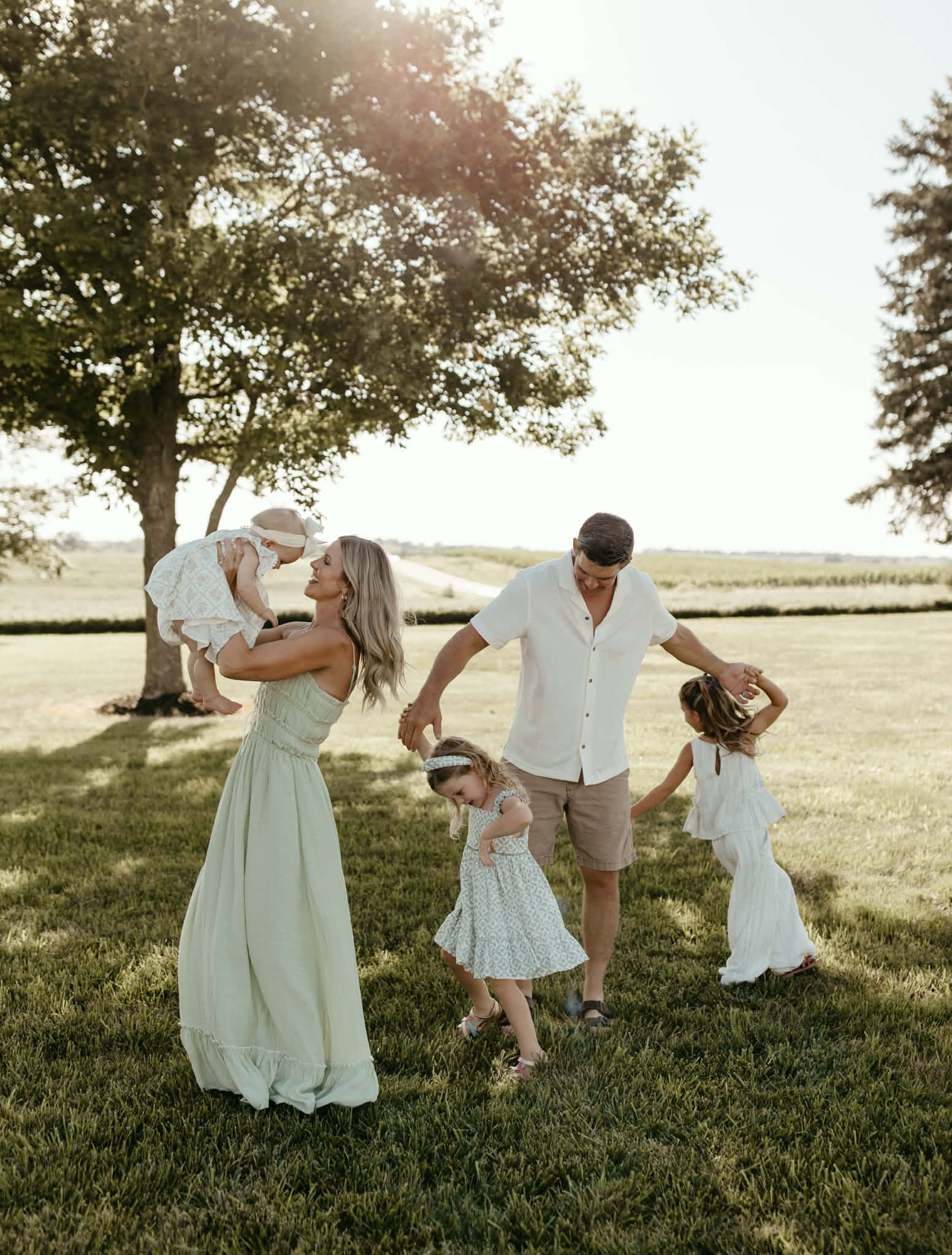 Family walking on grass in a sunny park, holding hands, with a small child in white dress and another being carried.